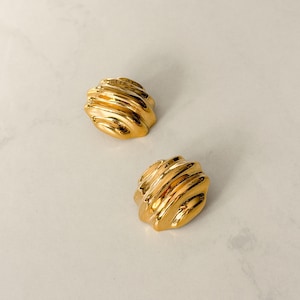 May include: Two gold-colored, textured stud earrings. Each earring has a rounded, ridged design, reflecting light. The earrings are displayed on a light-colored surface, showcasing their metallic sheen. These earrings are a classic jewelry accessory.