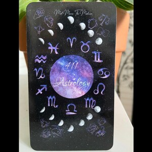 May include: A rectangular astrology card with a black background, featuring a central circle with the word "Astrology" and the number "411". The card is adorned with zodiac symbols, crescent moons, and the text "Mid Mane The Medium".