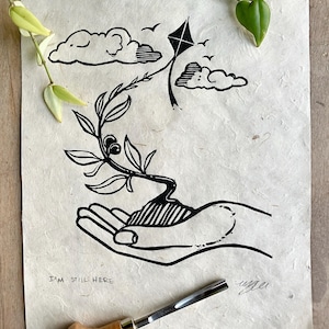 Op de afbeelding: Zwart-witte linosnede van een hand die een kleine plant vasthoudt met een vlieger die erboven vliegt. De print heeft de tekst "I'm still here" onderaan.