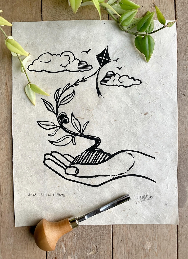 Op de afbeelding: Zwart-witte linosnede van een hand die een kleine plant vasthoudt met een vlieger die erboven vliegt. De print heeft de tekst "I'm still here" onderaan.