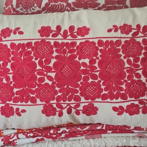 May include: A rectangular white pillow with vibrant red floral embroidery. The embroidery features large, stylized flowers and leaves, creating a bold pattern. The pillow is part of a set, with other embroidered items visible.