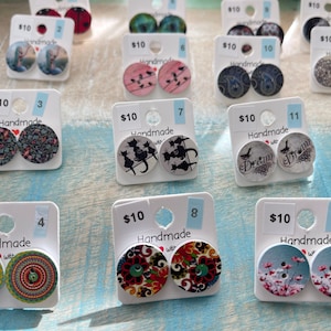 Peut inclure: Douze paires de boucles d'oreilles en bois faites à la main avec différents motifs, notamment des motifs floraux, des oiseaux, des chats et des motifs abstraits. Chaque paire coûte 10 € et est numérotée.