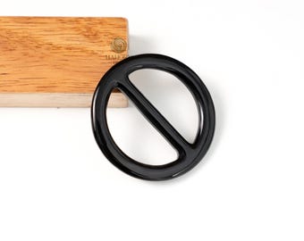 May include: A glossy black circular buckle with a diagonal bar across the center. The buckle is set against a white background with a wooden block in the upper left corner. The buckle is a fashion accessory.