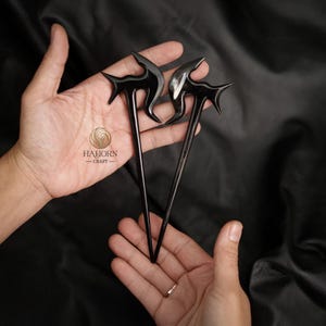 May include: Two black hair sticks with a unique, abstract design. The sticks are made of horn and have a polished finish.