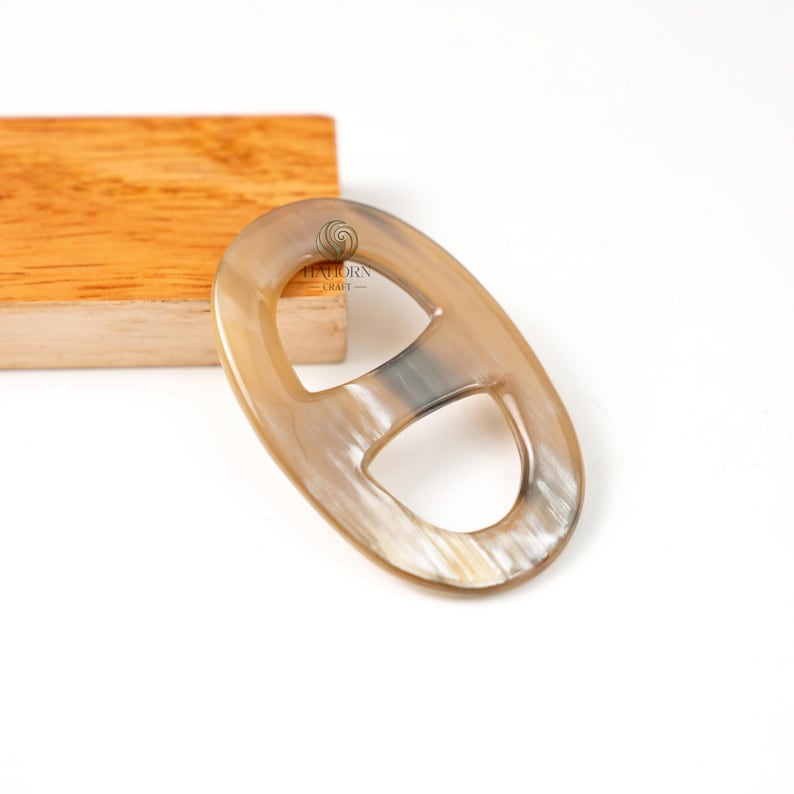 May include: Oval-shaped, horn-colored buckle with a central bar. The buckle has a natural, marbled appearance with shades of tan and cream. The product is set against a white background with a wooden block in the background.