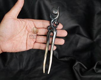 May include: A black and white hair stick with a carved design of a cat. The stick is about 6 inches long and has a two-pronged base.