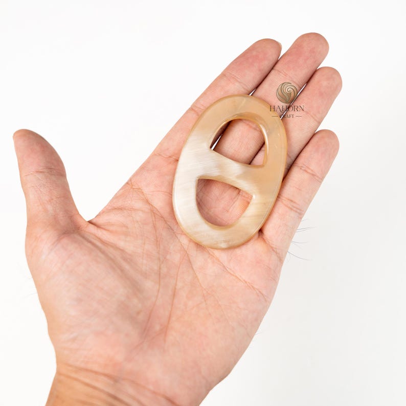 May include: A light beige oval-shaped accessory with a distinctive D-shaped cutout. The accessory has a smooth, polished surface and is held in a person's hand. The item is likely a decorative element or a fashion accessory.