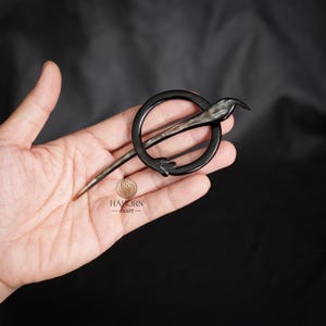 May include: A black horn hair stick with a circular design. The stick is approximately 15 cm long and features a bird-like design at the top. The stick is held in a hand.