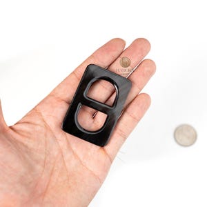 Puede incluir: Una hebilla negra rectangular con un recorte en forma de 'D' redondeada. La hebilla tiene un acabado brillante y se sostiene en una mano. El logotipo "HAJOR CRAFT" es visible en la esquina superior derecha. Una moneda está en el fondo.