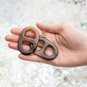 Op de afbeelding: Twee ovaalvormige, donkerbruine decoratieve sluitingen met een centrale balk, vastgehouden in een hand. De sluitingen hebben een glanzende afwerking en een gemarmerd uiterlijk. De achtergrond is een licht, getextureerd oppervlak.