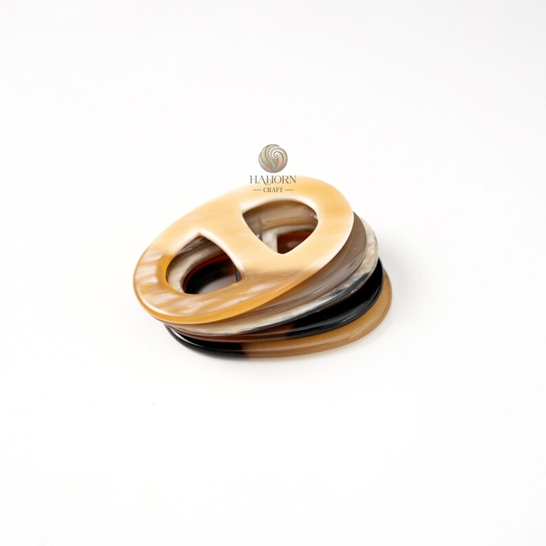 May include: A stack of oval-shaped, horn-like buckles in various colors, including tan, brown, and black. The buckles have a distinctive design with a central opening. The logo "HAJORN - CRAFT" is visible in the background.