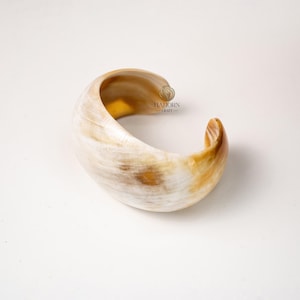 May include: A curved, open-ended cuff bracelet made of natural horn. The bracelet features a blend of creamy white and warm brown tones, with a polished, smooth surface. The overall design is organic and minimalist.