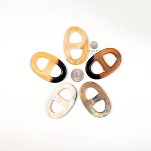 May include: Five oval-shaped horn links in varying shades of tan, brown, and black, arranged in a starburst pattern. Each link features a rectangular cutout in the center. A quarter is placed in the center of the links. The background is white.