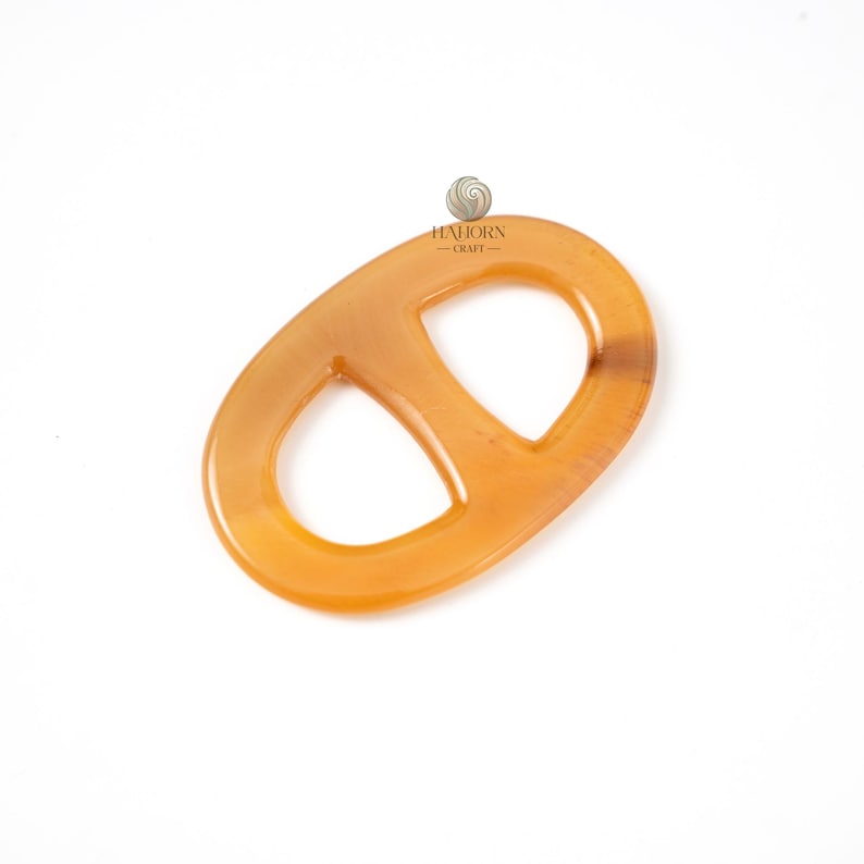 May include: A light amber-colored, oval-shaped accessory with a double-loop design. The accessory is made of a translucent material, possibly horn or resin, and has a smooth, polished surface. The item is set against a white background.