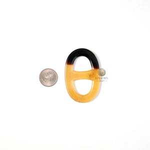 May include: A decorative, oval-shaped pendant with a gradient design. The top portion is black, transitioning to a light amber color. A US quarter is next to the pendant for size comparison. The pendant has a glossy finish.