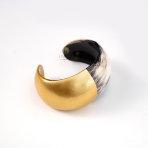 May include: A gold-colored cuff bracelet with a section of black and white patterned material. The bracelet has a curved design and is set against a white background. The gold section has a metallic sheen.