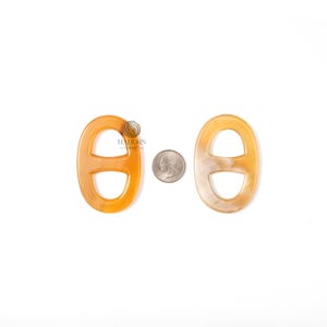 May include: Two amber-colored, oval-shaped chain link pendants with a quarter coin for size comparison. The pendants have a glossy finish and a classic design. The background is white.