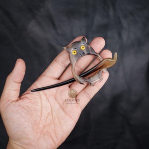 May include: A handcrafted owl-shaped hair barrette. The barrette features a circular design with an owl head and a long, black pin. The owl's eyes are yellow. The barrette is made of a light brown material with darker brown streaks.