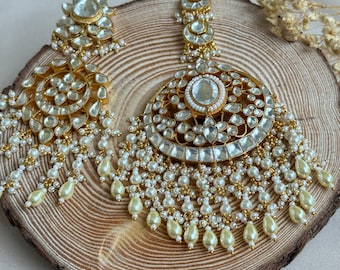 May include: Gold-toned, ornate earrings with clear, faceted stones and dangling pearl accents. The earrings feature a floral design and are displayed on a wooden surface. The jewelry has a traditional, elegant aesthetic.