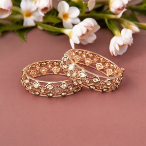 May include: Two gold bangles with intricate designs featuring small white stones and red accents.