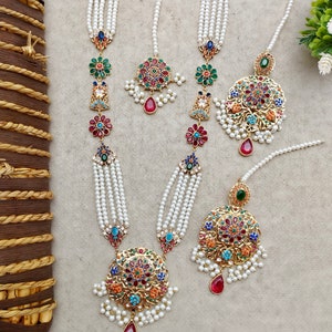 Op de afbeelding: Drie sets parel- en gouden kettingen met kleurrijke bloem- en juwelenhangers. Elke ketting heeft een grote ronde hanger met een rood, blauw, groen en gouden ontwerp.