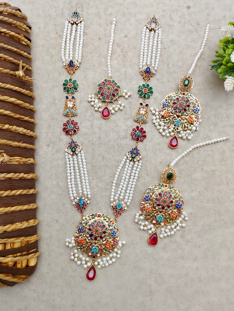 Op de afbeelding: Drie sets parel- en gouden kettingen met kleurrijke bloem- en juwelenhangers. Elke ketting heeft een grote ronde hanger met een rood, blauw, groen en gouden ontwerp.