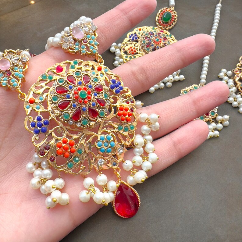 Op de afbeelding: Een gouden ketting met een grote, sierlijke hanger met een bloemmotief met gekleurde stenen. De hanger is omringd door een rij witte parels.
