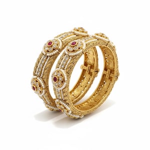 May include: Two gold bangles with intricate detailing. Each bangle features red gemstones, small white pearls, and clear crystals. The bangles have a hinged design and are set against a white background.