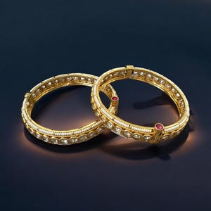 May include: Two gold bangles with intricate detailing. Each bangle features a row of clear stones, white beads, and a small red stone. The bangles are set against a dark blue background, highlighting their golden hue.