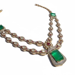 May include: A gold-toned necklace featuring a large, rectangular emerald pendant. The necklace has two strands of smaller gemstones, including emeralds, rubies, and clear stones. The design is intricate and ornate, with a focus on luxury and elegance.