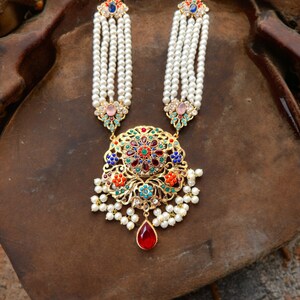 Op de afbeelding: Een gouden ketting met een grote, sierlijke hanger met een kleurrijk bloemmotief. De hanger is omringd door witte parels en hangt aan een streng witte parels.