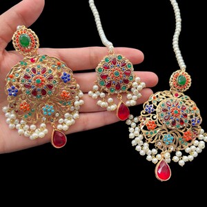 Op de afbeelding: Een set gouden oorbellen en een maang tikka met rode, groene, blauwe en oranje edelstenen. De oorbellen en maang tikka zijn versierd met witte parels.