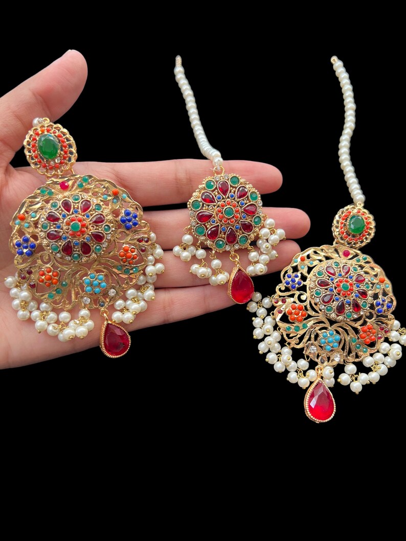 Op de afbeelding: Een set gouden oorbellen en een maang tikka met rode, groene, blauwe en oranje edelstenen. De oorbellen en maang tikka zijn versierd met witte parels.