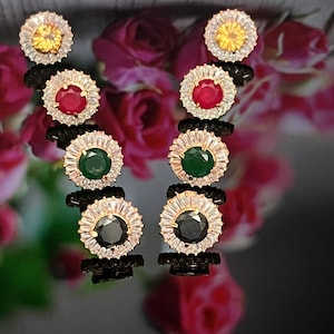 May include: A pair of stud earrings with a gold setting. Each earring features a central gemstone in yellow, red, green, and black, surrounded by a halo of clear, baguette-cut stones. The earrings are displayed against a blurred floral background.