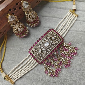 May include: A gold choker necklace with a rectangular pendant set with clear and pink stones. The necklace is made of multiple strands of white pearls and has pink and clear dangling charms. The necklace comes with a pair of matching earrings.
