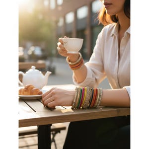 May include: A collection of colorful bangles adorns a wrist, with a variety of hues including red, green, yellow, and gold. The bangles are stacked together, creating a vibrant and eye-catching display. A white teacup is held in the other hand.
