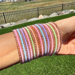May include: A selection of pastel-coloured bangles on a wrist. The bangles are in shades of pink, blue, green, and orange, with a gold-coloured beaded trim. The background shows a green lawn and a residential area.
