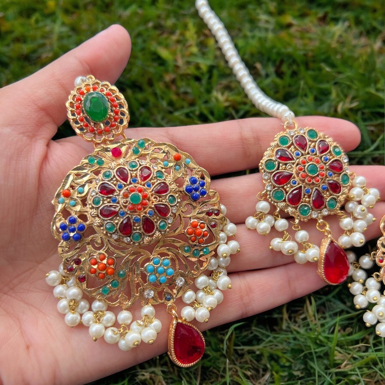 Op de afbeelding: Een paar gouden oorbellen met rode, groene, blauwe en oranje edelstenen. De oorbellen zijn versierd met witte parels en hebben een traanvormige hanger.