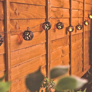 May include: A string of decorative, spherical outdoor lights with a warm glow. The black lights have intricate cut-out patterns, hanging against a wooden fence. The fence is made of horizontal wooden planks, and the background is blurred.