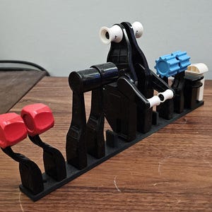 Op de afbeelding: Een zwart 3D-geprint model met rode, witte en blauwe accenten. Het model heeft verschillende componenten, waaronder twee rode dobbelstenen, een centrale structuur met witte cilinders en een blauw tandwielachtig stuk. Het staat op een houten oppervlak.