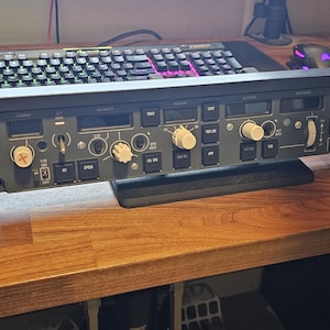 Op de afbeelding: Een zwart en grijs bedieningspaneel voor een vluchtsimulator met talrijke knoppen, toetsen en schakelaars. Het paneel staat op een houten bureau, met een toetsenbord en andere computeraccessoires op de achtergrond. Het paneel heeft een zwarte basis en witte knoppen.