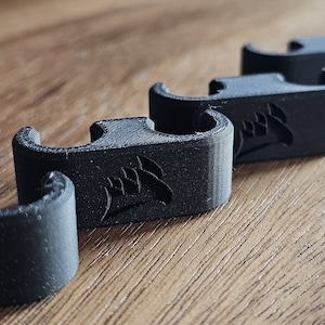 Könnte beinhalten: Fünf schwarze Kunststoff-Kabelclips mit einem Logo auf jedem. Die Clips sind so konzipiert, dass sie Kabel an Ort und Stelle halten.