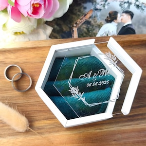 May include: A hexagonal white ring box with a clear acrylic lid featuring white etched botanical frames, the initials A & W, and the date 06.06.2026. The interior is lined with deep teal velvet, and two plain silver-toned metal bands sit nearby.