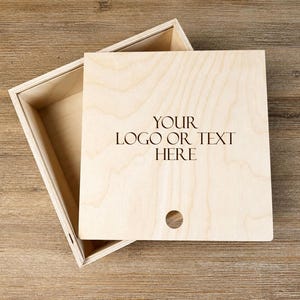 Könnte beinhalten: Eine hellfarbene Holzkiste mit Deckel. Der Deckel ist geöffnet und gibt das Innere der Kiste frei. Der Deckel trägt den Text "YOUR LOGO OR TEXT HERE" und ein kleines kreisförmiges Loch. Die Kiste steht auf einer Holzoberfläche.