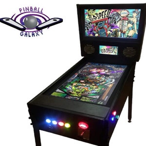 May include: Black pinball machine with a colorful playfield and a screen displaying a car-themed graphic. The machine has a "Pinball Galaxy" logo and a "Vs Speed" graphic. It features multi-colored lights and a "Launch Ball" button.