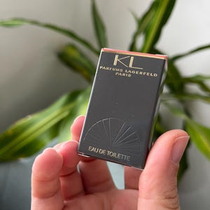 Peut inclure: Boîte de parfum rectangulaire noire avec l'inscription dorée "KL PARFUMS LAGERFELD PARIS". Le bas de la boîte présente un motif en éventail et l'inscription "EAU DE TOILETTE". Le dessus de la boîte est saumon.
