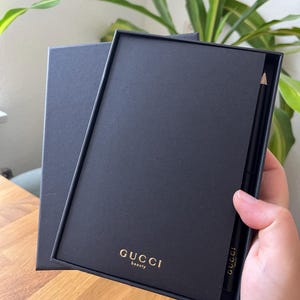 May include: A black box with the gold text "GUCCI beauty" is open to reveal a black pencil. The pencil has the word "GUCCI" printed on it. The box is on a wooden surface.