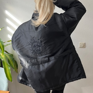 Puede incluir: Chaqueta negra con el logo de Versace Jeans Couture bordado en la espalda. La chaqueta tiene un cuello alto y una cintura ajustable. El logo presenta un diseño circular con el nombre de la marca y una cabeza de Medusa.