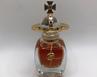 Op de afbeelding: Een heldere glazen fles met een gouden bolvormige dop en een gouden kruis bovenop. De fles bevat een bruine vloeistof en heeft een gouden etiket met een symbool.
