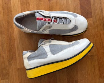 May include: A pair of low-top trainers featuring white patent leather overlays, grey mesh panels, and grey laces. The tongue displays a red label with the white text PRADA. The shoes have a thick, bright yellow midsole and a black outsole.